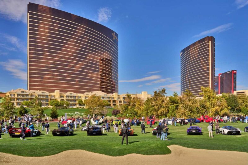 A large crowd gathers on a grassy area displaying luxury cars at the 2025 Las Vegas Concours Becomes Hypercar Ground Zero, with two tall, reflective Wynn hotel towers in the background under a clear blue sky.
