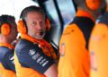 A man in an orange and navy team uniform wearing headphones and a microphone sits at a console, looking toward the camera—perhaps inspired by insights from McLaren Racing CEO Zak Brown Reveals Secrets To Success In New Autobiography. Two teammates are beside him.