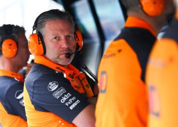 A man in an orange and navy team uniform wearing headphones and a microphone sits at a console, looking toward the camera—perhaps inspired by insights from McLaren Racing CEO Zak Brown Reveals Secrets To Success In New Autobiography. Two teammates are beside him.