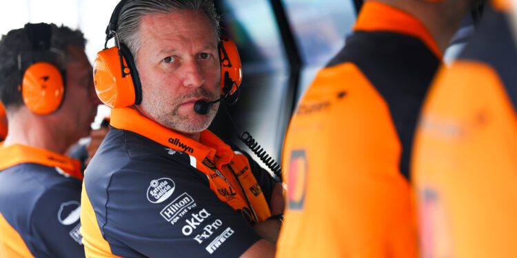 A man in an orange and navy team uniform wearing headphones and a microphone sits at a console, looking toward the camera—perhaps inspired by insights from McLaren Racing CEO Zak Brown Reveals Secrets To Success In New Autobiography. Two teammates are beside him.