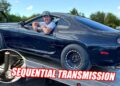 A man leans out of the window of a dark Toyota Supra sports car on a trailer; inset shows a Sequential Gearbox shifter. Text reads "SEQUENTIAL TRANSMISSION.
