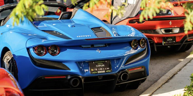 A blue Ferrari convertible and a red Ferrari coupe are parked side by side at an outdoor Cars on 5th event, surrounded by greenery and people enjoying the luxury automotive experience.