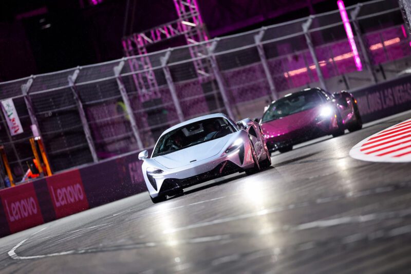 Two sports cars are driving around a corner on the Las Vegas F1 track at night, with barriers and pink lighting illuminating the background.