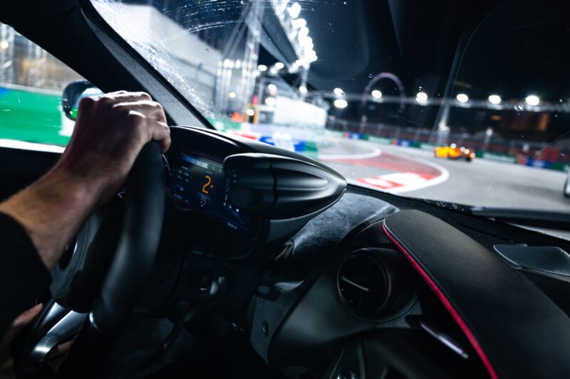 A driver steers a car on the brightly lit Las Vegas F1 Track at night, with another race car visible ahead through the windshield.