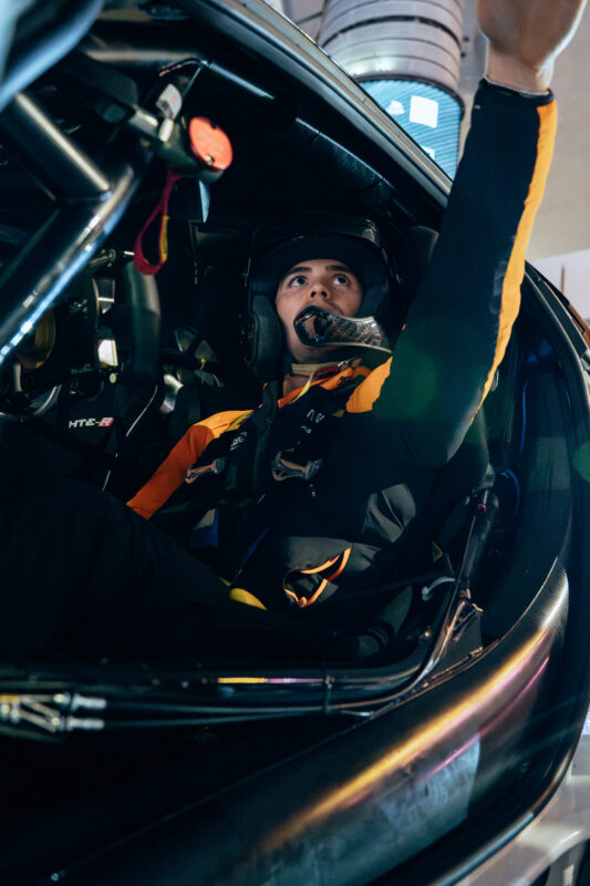 A McLaren race car driver wearing a helmet and racing suit sits inside a car cockpit at the Las Vegas F1 Track, looking up and reaching one arm toward the roof.