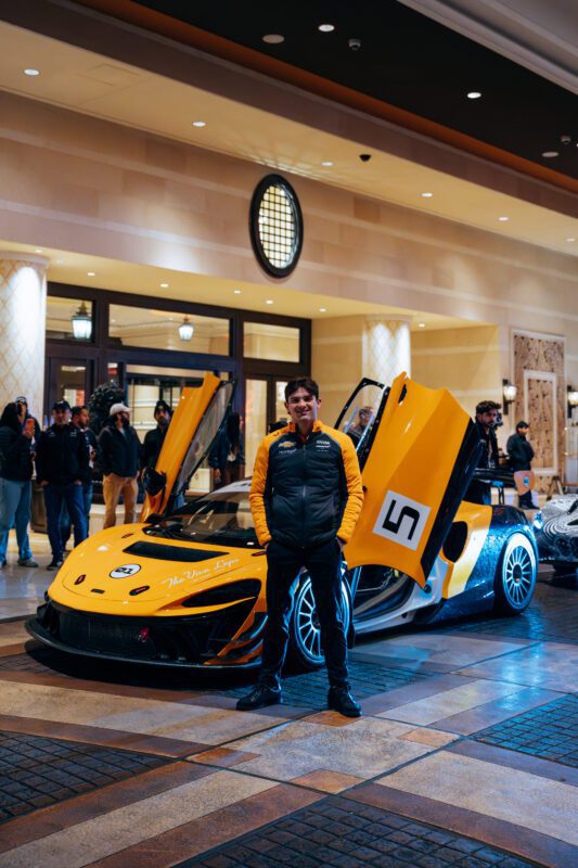 A man stands in front of a yellow and black McLaren sports car with its doors open, inside a well-lit building, with several people in the background.