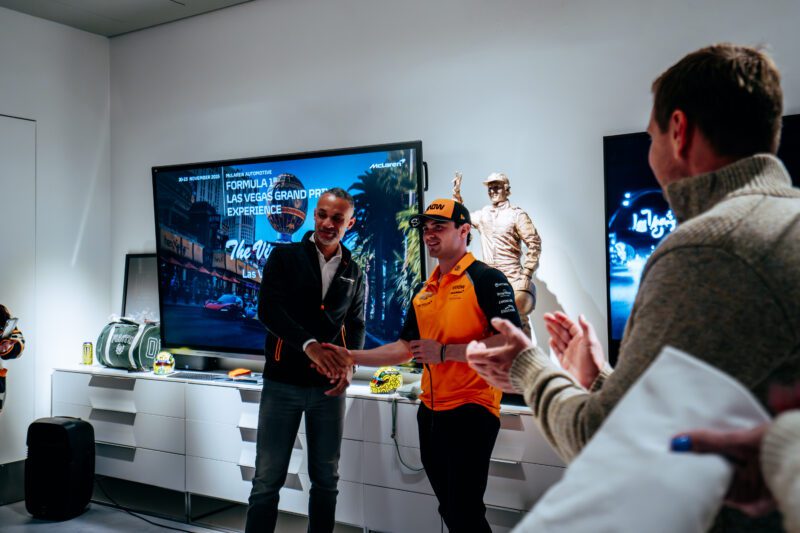 Two men shake hands in front of a TV displaying “Formula 1 Las Vegas Grand Prix Experience” at the brightly lit Las Vegas F1 Track, as people applaud enthusiastically.