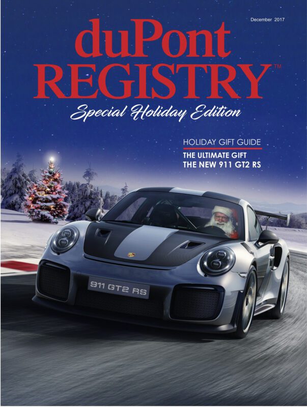 A silver Porsche 911 GT2 RS, featured in duPont REGISTRY’s Cover Cars for the 40th Anniversary, cruises on a snowy road with Santa Claus behind the wheel. A decorated Christmas tree and snow-covered trees add festive flair to the background.