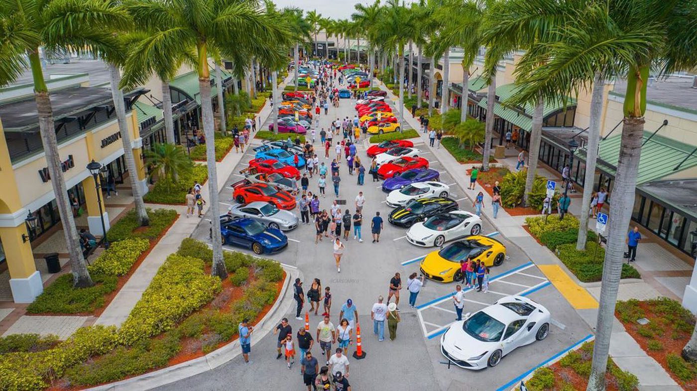 A large crowd walks among rows of colorful luxury sports cars, including a rare Praga Bohema, displayed along a palm-lined outdoor shopping plaza at this South Florida automotive event.