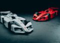 Two Porsche Formula E factory cars, one white and one red, are parked on a dark, textured surface in a dimly lit environment.