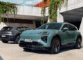 Two modern electric SUVs, including The Most Powerful Production Porsche Ever: 2026 Cayenne Electric, are parked outside a concrete building with plants hanging from above; both vehicles have European license plates.