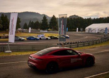 A red Porsche is parked on a racetrack near a curve, with several cars lined up on the other side and banners and trees in the background.