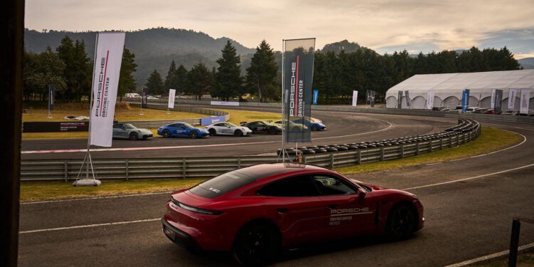 A red Porsche is parked on a racetrack near a curve, with several cars lined up on the other side and banners and trees in the background.