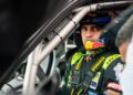 A race car driver wearing a helmet and racing suit sits inside a car, looking out through the window, with safety equipment visible.