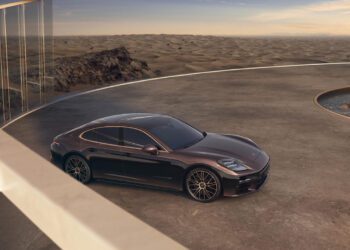 A dark-colored luxury sedan is parked on a circular concrete platform with a glass railing, overlooking a desert landscape at sunset.
