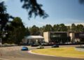 Several sports cars navigate a race track near a modern building labeled "Porsche Driving Center Helsinki," with trees and clear sky in the background.