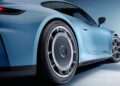 Close-up of the rear side of a blue Porsche sports car, highlighting the wheel, tire, and aerodynamic body design against a minimal, light blue background.