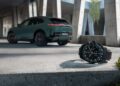 A wristwatch from ICustom Timepieces with a black strap rests on a concrete surface in the foreground, while a green SUV from the Porsche Design Cayenne Models is parked in the background under a modern structure.