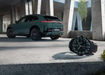 A wristwatch from ICustom Timepieces with a black strap rests on a concrete surface in the foreground, while a green SUV from the Porsche Design Cayenne Models is parked in the background under a modern structure.
