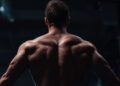 A muscular man is shown from behind in a dimly lit gym, highlighting his well-defined back and shoulder muscles—an inspiring image of energy and dedication to longevity.