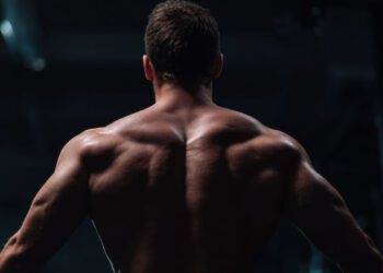 A muscular man is shown from behind in a dimly lit gym, highlighting his well-defined back and shoulder muscles—an inspiring image of energy and dedication to longevity.