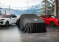 Two Porsche SUVs, a white and a red Cayenne Electric, flank a third vehicle hidden by a black cloth in a Dubai showroom with large windows and mountain views.