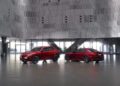 Two red Alfa Romeo vehicles, an SUV and a sedan, are parked inside a modern building with metallic perforated walls and large windows.