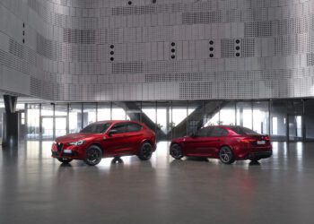 Two red Alfa Romeo vehicles, an SUV and a sedan, are parked inside a modern building with metallic perforated walls and large windows.