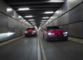 Two red Alfa Romeo cars drive side by side through a modern, well-lit tunnel with metallic walls.