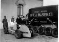 Three men stand beside a vintage Maserati race car, a truck with "Società Anonima Off. A. Maserati" branding, and several spare tires outdoors.