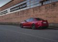 A red Alfa Romeo Giulia Quadrifoglio is parked on a street beside a long brick industrial building with large windows.