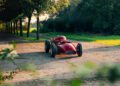 A vintage red race car is parked on a sunlit path surrounded by trees and greenery.