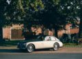 A vintage silver Maserati coupe is parked on a street in front of trees, a brick building, and various farm equipment and animal sculptures—capturing timeless style for the Maserati 111th Anniversary.