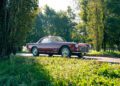 A classic red Maserati coupe car parked on a sunlit road, surrounded by green grass and tall trees in a park-like setting, celebrating the 111th Anniversary of Maserati.