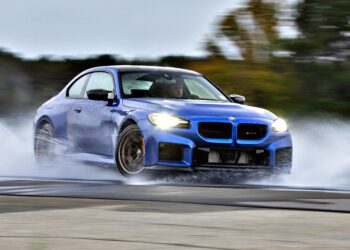 A blue 2026 BMW M2 CS, the lightest M car you can buy new, speeds through a wet track, creating large splashes of water from its tires, with blurred trees in the background.