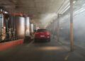 A red Alfa Romeo Stelvio Quadrifoglio is parked inside an industrial warehouse with large tanks on the left and light streaming in from windows on the right.
