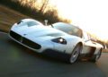 A white and blue Maserati sports car drives on a paved road at sunset, celebrating the brand’s 111th Anniversary amid trees and a blurred landscape in the background.