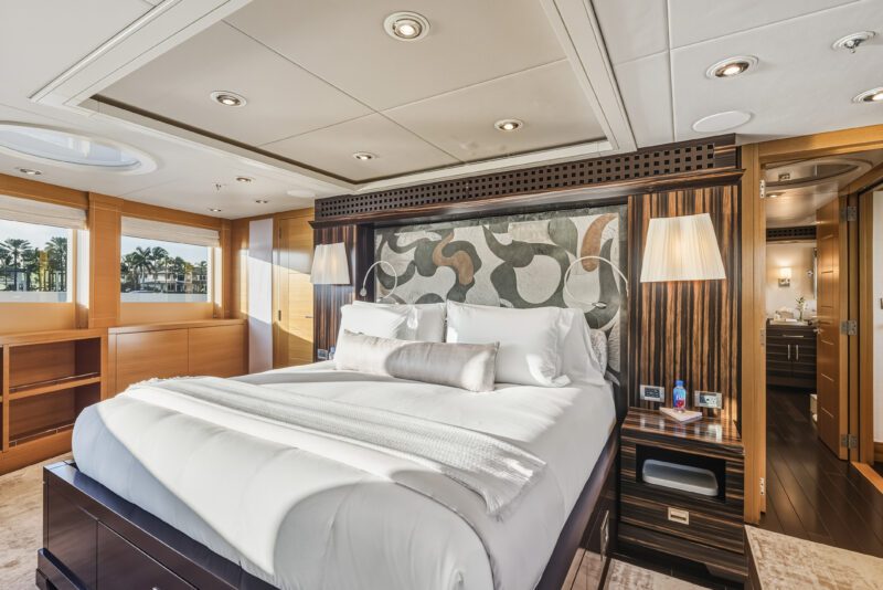 Modern yacht bedroom with a large bed, abstract headboard, pendant lamps, wood accents, and a water view through windows—an inviting retreat aboard the five-star yacht Delta Marine Noverra.