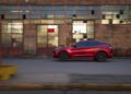 A red Alfa Romeo Stelvio Quadrifoglio SUV is driving past an old brick building with large windows and industrial doors in daylight.