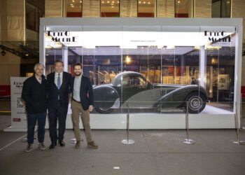 Three men stand before a glass case featuring a 1937 Bugatti Type 57S Atalante at the Rétromobile New York outdoor exhibition, where ticket sales are brisk.