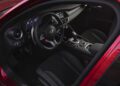 View of the driver’s seat and dashboard of a modern Alfa Romeo with black interior, digital displays, and red stitching on the seats and steering wheel.