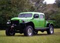 Bright green vintage Dodge Power Wagon truck with large off-road tires parked on grass, trees in the background.