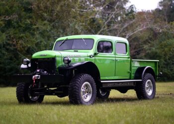 Bright green vintage Dodge Power Wagon truck with large off-road tires parked on grass, trees in the background.