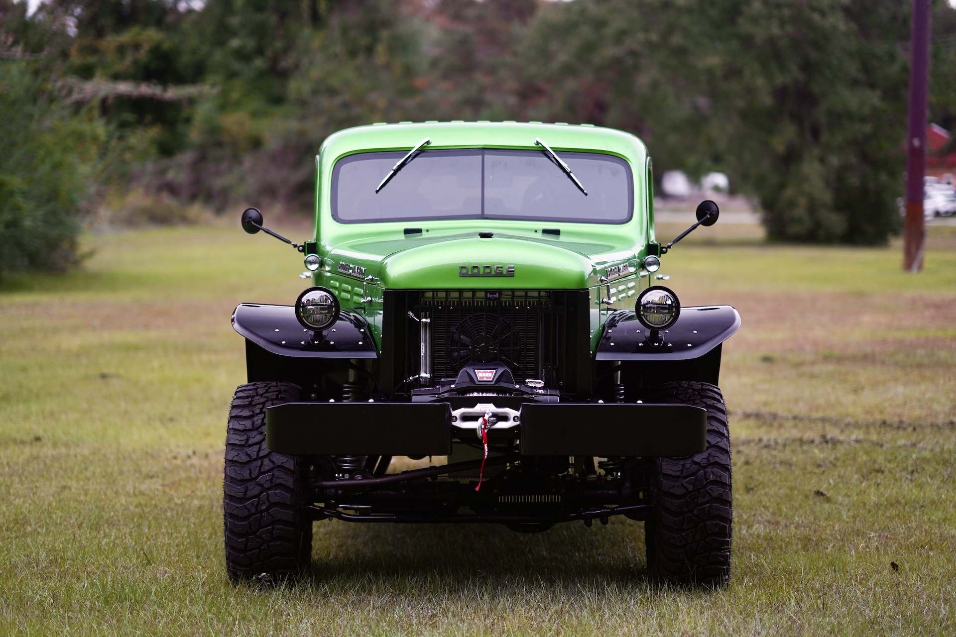 Front view of a vintage, bright green Dodge Power Wagon parked on grass, with large tires and round headlights, set outdoors with trees in the background.