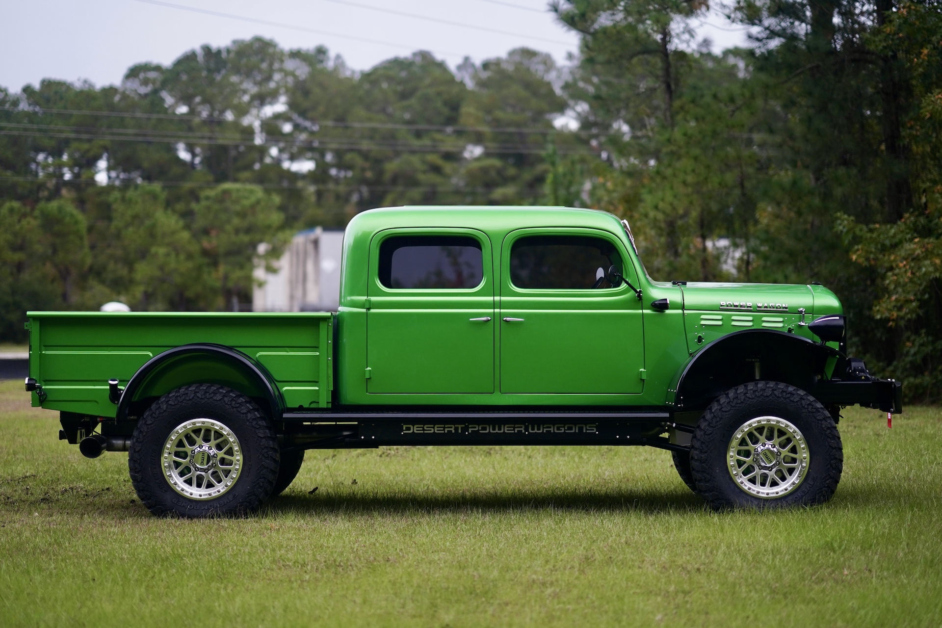 A bright green vintage Dodge Power Wagon Crew Cab with four doors and large off-road tires is parked on grass, with trees and power lines in the background.