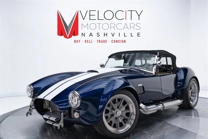 A blue Shelby Cobra with white racing stripes is displayed in a showroom with a Velocity Motorcars Nashville sign in the background, highlighting their impressive selection of classic muscle cars and high-performance cars.