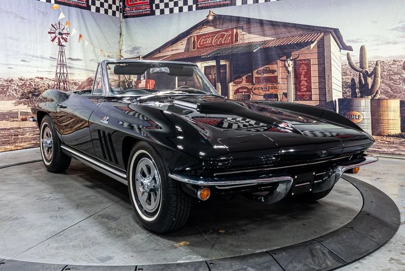 A classic black convertible car is displayed indoors with a vintage gas station backdrop, featuring old signs and a Coca-Cola advertisement—an iconic scene for fans of classic muscle cars.