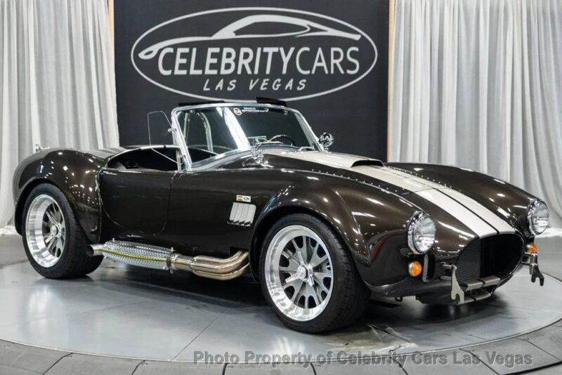A black classic convertible sports car with white racing stripes is displayed indoors on a rotating platform at Celebrity Cars Las Vegas, showcasing one of the finest classic muscle cars in their collection.