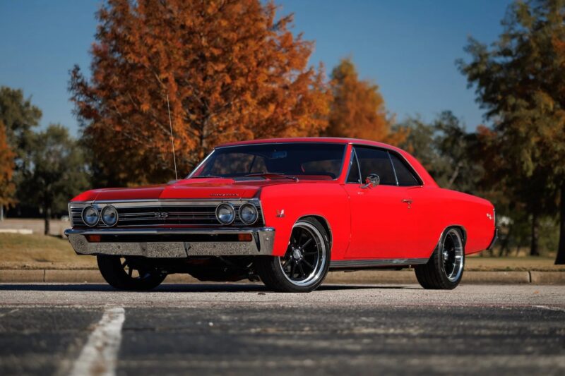 A red classic muscle car is parked on asphalt in front of autumn trees under a clear blue sky, capturing the timeless appeal of Classic Muscle Cars.