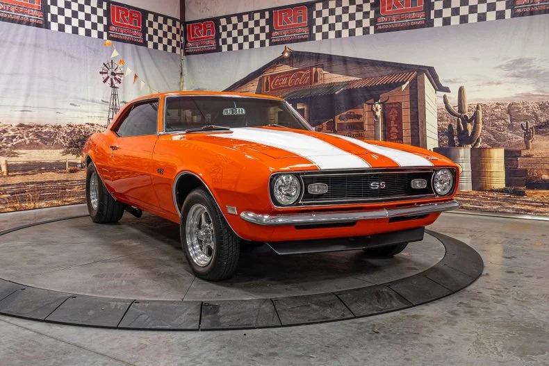 An orange 1968 Chevrolet Camaro SS with white racing stripes is displayed indoors on a circular platform, embodying the spirit of classic muscle cars against a striking Western-themed backdrop.